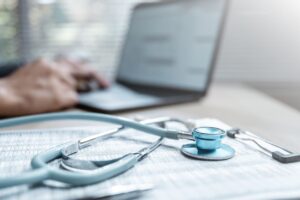 Stethoskop liegt auf medizinischen Unterlagen, während im Hintergrund eine Person an einem Laptop arbeitet